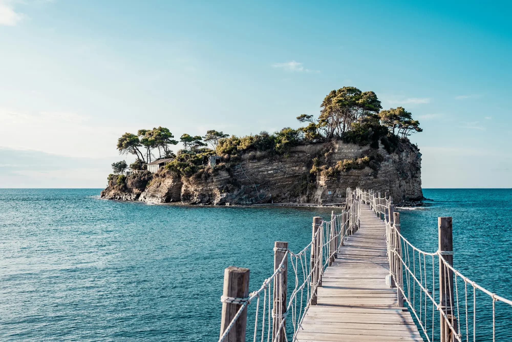 A wooden rope bridge leading across turquoise water to a small rocky island topped with pine trees